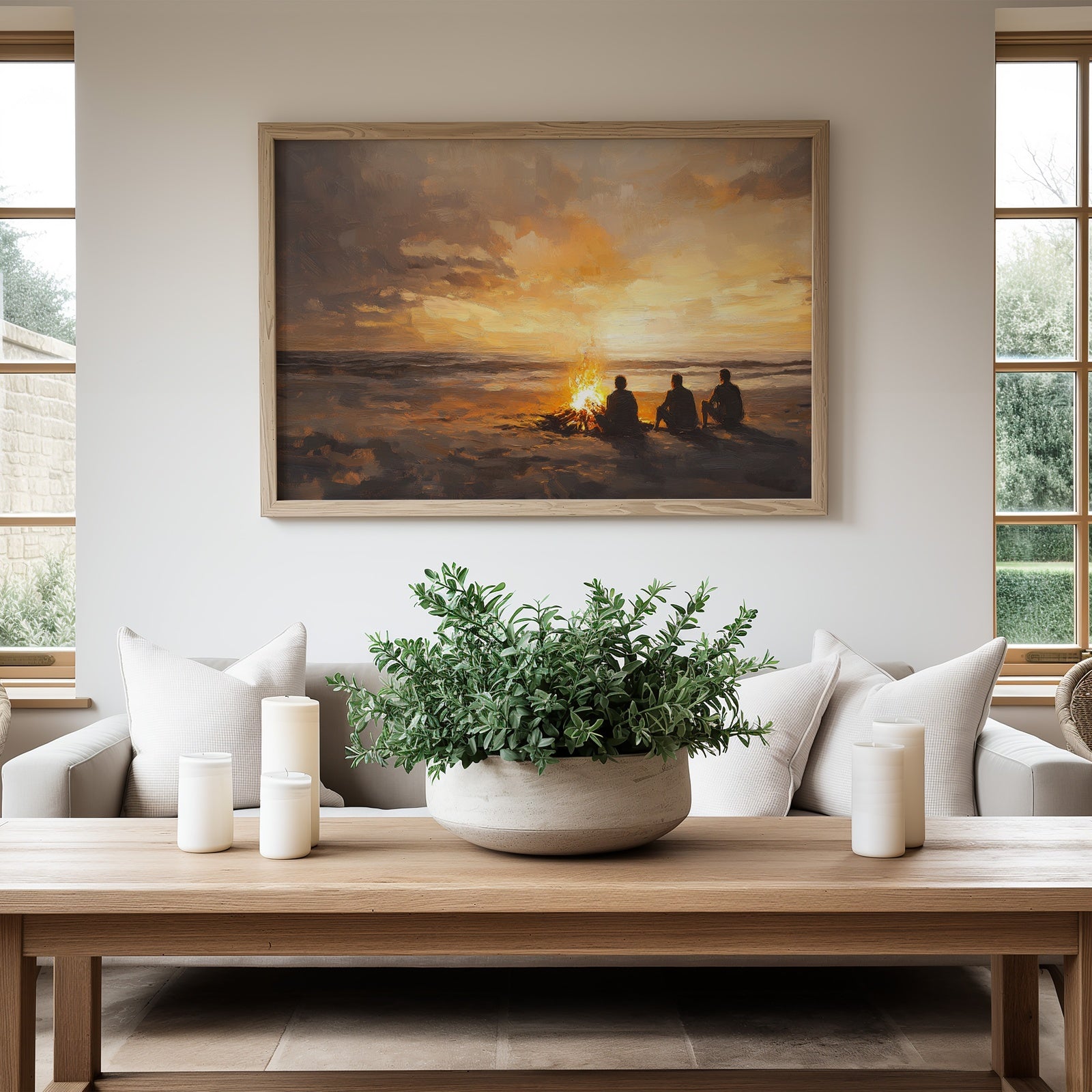 A modern living room with a framed digital art print depicting three people sitting by a bonfire on the beach at sunset. The warm golden hues and painterly style create a cozy coastal atmosphere, blending perfectly with the neutral and natural decor
